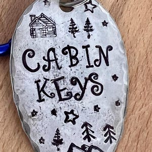 May include: A silver metal cabin key tag with a blue carabiner clip. The tag is stamped with the words "CABIN KEYS", a cabin illustration, trees, stars, and mountains.