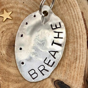 May include: A silver key chain with a round tag that says "BREATHE" in a hammered metal style.