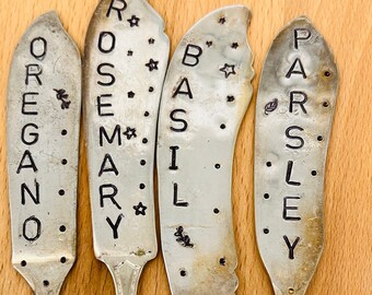 May include: Four vintage silver herb garden markers. Each marker is a repurposed spoon or knife, stamped with the names "OREGANO", "ROSEMARY", "BASIL", and "PARSLEY". Decorative star and leaf accents are also stamped on each marker.