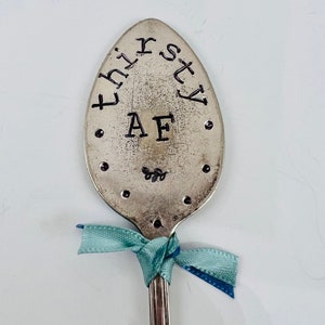 May include: A silver spoon with the words "thirsty AF" stamped on the spoon's head. The spoon is decorated with small black dots and a teal ribbon tied around the handle.