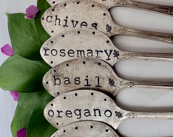 May include: Six vintage silver spoons repurposed as herb garden markers. Each spoon is stamped with a different herb name: dill, chives, rosemary, basil, oregano, and thyme. The spoons are arranged with green leaves and purple flower petals.