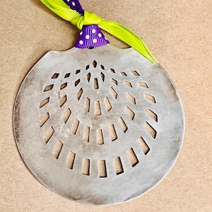 May include: A silver-colored, decorative ornament with a circular design featuring rectangular cutouts. It is adorned with a purple ribbon with white polka dots and a bright green ribbon, tied in a knot at the top.
