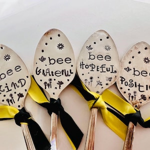 May include: Four silver spoons with black lettering that reads "bee kind", "bee grateful", "bee hopeful", and "bee positive". The spoons are tied together with black and yellow ribbon.