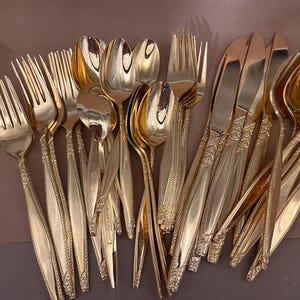 May include: A collection of gold-colored flatware, including forks, spoons, and knives. The utensils have a decorative pattern on the handles and are arranged on a brown surface. The silverware is likely for a formal dining setting.