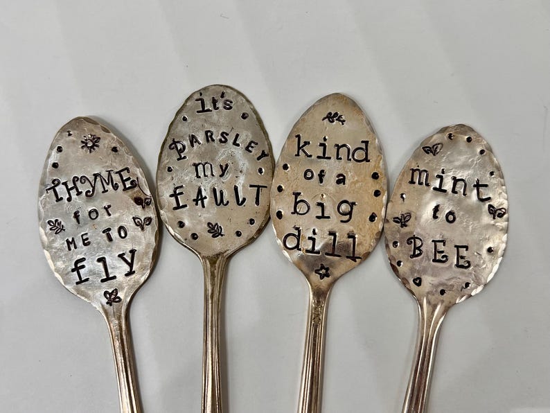 May include: Four silver spoons with engraved sayings about herbs. The spoons read "Thyme for me to fly", "It's Parsley my fault", "Kind of a big dill", and "Mint to be".