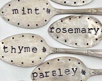 May include: Six silver spoons with black lettering, each labeled with a different herb: dill, mint, rosemary, thyme, parsley, and oregano. Each spoon has a small leaf design next to the herb name.