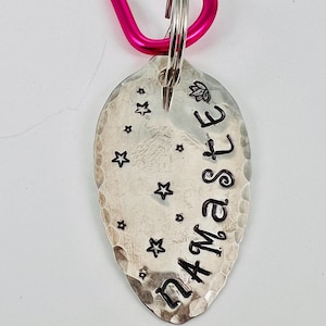 May include: A silver spoon keychain with the word "Namaste" engraved on it. The spoon has a hammered texture and is decorated with small stars. The keychain is attached to a pink carabiner.