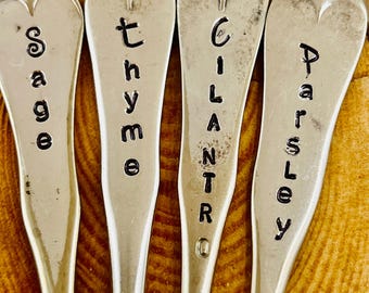 May include: Four silver herb markers with the names "Sage", "Thyme", "Cilantro", and "Parsley" stamped in black. The markers have a spoon-like shape with a heart-shaped top. The background is a wooden surface.