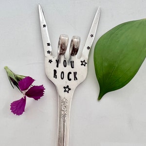 May include: A silver fork with the words "YOU ROCK" stamped on it. The fork is bent into a rock and roll hand gesture. There are small stars stamped around the words and the fork tines. A green leaf and a purple flower are in the background.