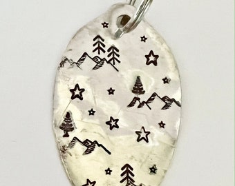 May include: A silver oval keychain with a design of mountains, trees, and stars. The keychain has a silver ring attached to the top.