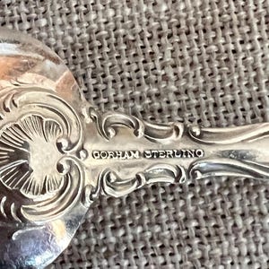 May include: Close-up of a silver spoon with ornate floral and scrollwork detailing. The spoon's handle is marked "GORHAM STERLING" in a classic serif font. The spoon is set against a textured, neutral-toned background.