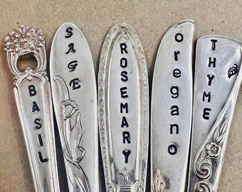 May include: Five vintage silver spoons repurposed as herb markers. Each spoon is engraved with a different herb name: Basil, Sage, Rosemary, Oregano, and Thyme. The spoons have decorative handles and are arranged against a neutral background.