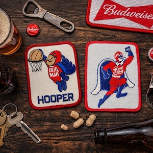 May include: An overhead shot of a wooden table with beer-related items. Includes a glass of beer, a bottle, bottle openers, coasters with a superhero design, bottle caps, and keys. The coasters feature a superhero with a basketball and the word "HOOPER".