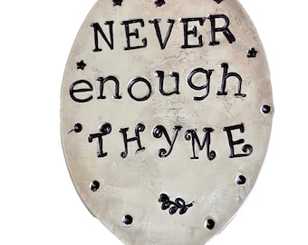 May include: A silver-colored spoon with the words "NEVER enough THYME" stamped on the spoon's head. The spoon is decorated with small star and leaf designs. The spoon is a kitchen utensil.
