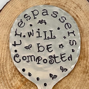May include: A silver spoon with the words "You will be composted" stamped on it. The spoon is sitting on a wooden surface.