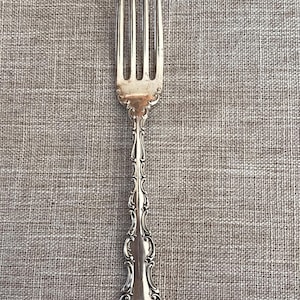 May include: A silver fork with four tines and an ornate handle. The handle features a detailed, scrolling design. The fork is set against a neutral, textured background, highlighting its elegant details. This is a vintage silverware piece.