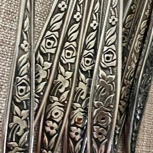May include: Close-up of vintage silverware with floral rose and leaf pattern. The silver utensils have a detailed design of roses and leaves. The silverware is arranged on a neutral-colored surface.