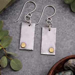May include: A pair of silver rectangular earrings with a hammered texture, each featuring a small, round gold accent near the bottom. The earrings hang from silver ear wires. The background includes greenery and a dark gray surface.