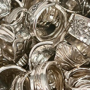 Może przedstawiać: Zbliżenie na stos srebrnych pierścionk&oacute;w wykonanych z przerobionych sztućc&oacute;w. Pierścionki są ułożone jeden na drugim i zachodzą na siebie, ukazując skomplikowane detale i wzory.