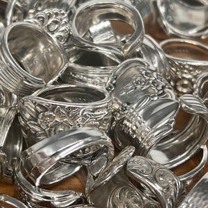 May include: A collection of handcrafted silver rings, each with unique designs and textures. The rings vary in style, some with floral patterns, others with ribbed or smooth bands. The rings are made from repurposed silverware.