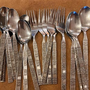 May include: A collection of vintage silverware, including spoons and forks, arranged on a brown surface. The silverware has a decorative square and swirl pattern on the handles. The spoons and forks are made of a shiny, silver-colored metal.