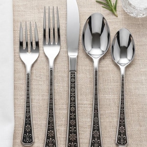 May include: A five-piece stainless steel flatware set consisting of a salad fork, dinner fork, dinner knife, dinner spoon, and teaspoon. Each handle features a dark, recessed decorative pattern of repeating floral and fleur-de-lis motifs.