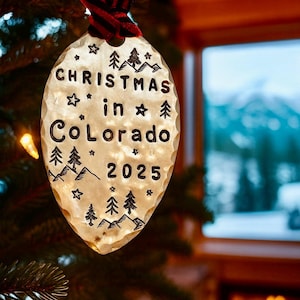 May include: A Christmas ornament with the text "Christmas in Colorado 2025" etched on a hammered metal oval. The ornament is adorned with mountain and tree designs, and hangs from a red and black striped ribbon.