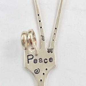 May include: Silver peace sign charm with the word "Peace" engraved on it. The charm is shaped like a hand making the peace sign. There are two rings attached to the charm.