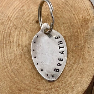 May include: A silver-colored metal keychain with the word "BREATHE" stamped on it. The keychain is oval-shaped with a series of small dots around the edge. A silver key ring is attached to the top. The background is a wooden surface.