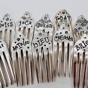 May include: A set of 10 silver forks with different names engraved on them, including "Goat", "As I Go", "Bleu", "Cheddar", "Havarti", "Brie", "GDAT", "Goldia", "Jack", and "Swiss".