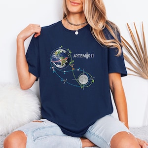 May include: Navy blue t-shirt featuring a graphic of Earth, the Moon, and a flight path, with the text "ARTEMIS II" in white. The design includes colorful dots and lines. The shirt is made of a soft material.