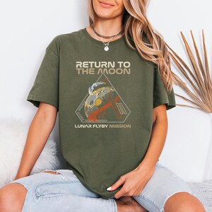 May include: Olive green t-shirt with the text "RETURN TO THE MOON" above a triangular graphic. The graphic features a stylized image of the moon and a rocket, with the text "LUNAR FLYBY MISSION" below.