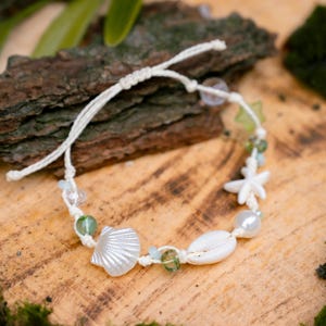 Puede incluir: Pulsera artesanal con temática de playa. Presenta una concha blanca, una estrella de mar y cuentas verdes y transparentes. Ensartada en un cordón crema, ideal para un look veraniego y con elementos náuticos.