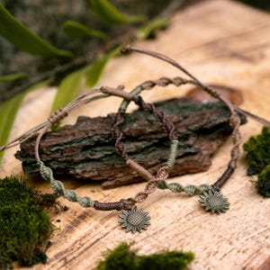 May include: Two handmade macrame bracelets with sunflower charms. The bracelets feature a braided design with shades of green, brown, and tan. The sunflower charms are a bronze color. The bracelets are displayed on a piece of wood.