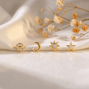 May include: Four gold-tone stud earrings featuring celestial motifs: a planet with a ring, a crescent moon with a small star, a sun with rays, and a five-pointed star. Each earring is accented with small, clear, round-cut white gemstones.