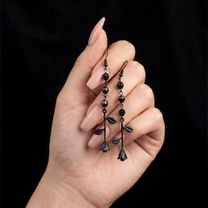 Puede incluir: Un par de pendientes largos con dijes de rosas de metal negro en la base, cada uno unido a un tallo con tres hojas pequeñas y una fila vertical de tres cuentas negras facetadas, terminados con ganchos de metal negro para orejas perforadas.