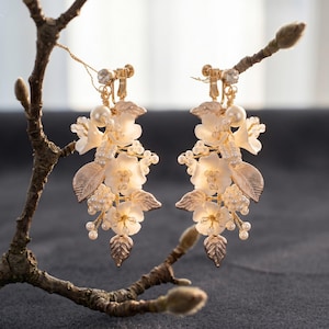 May include: A pair of gold-toned clip-on earrings featuring white floral blossoms, metallic leaves, and small white pearl-like beads. The design is arranged in a cascading cluster, suspended from a clear crystal accent at the top of each earring.