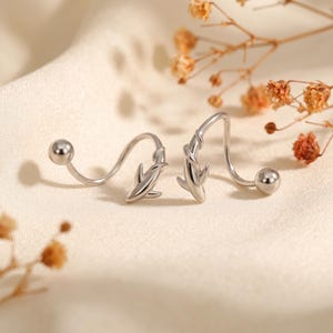 May include: A pair of silver-toned earrings featuring a curved, spiral-shaped wire design. Each earring incorporates a small, stylized shark figure integrated into the metal curve, finished with a smooth, polished ball at the end of the wire.