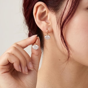 May include: A pair of gold hoop earrings featuring oval-shaped clear faceted gemstones. One earring is worn on the earlobe, while the other is held between the thumb and index finger, showcasing the reflective, brilliant cut of the stone.
