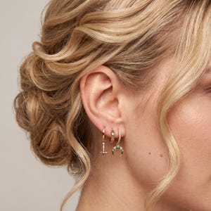 May include: Three gold hoop earrings worn in a single ear. The first hoop features a dangling star charm with a pink center stone. The second is a small hoop with multicolored stones. The third hoop features a crescent moon charm with green stones.