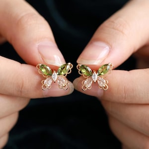 Pode incluir: Um par de brincos em forma de borboleta feitos de metal dourado. Cada brinco apresenta duas pedras verde-oliva em corte marquise para as asas superiores, duas pedras transparentes em corte de gota para as asas inferiores e uma pequena pedra central.