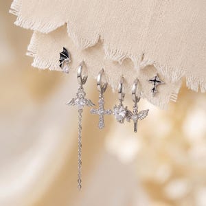 May include: A set of six silver-toned earrings featuring various designs including a black bat wing with a clear stone, a heart with wings and a long chain, a cross, a heart, a winged heart, and a black cross with clear stones.