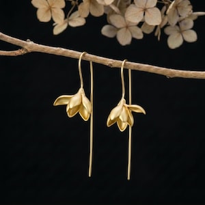 Op de afbeelding: Een paar goudkleurige oorbellen met een bloemknopontwerp. Elke oorbel bestaat uit een matte gouden bloemvorm die hangt aan een lange, dunne, rechte gouden draadhaak, wat zorgt voor een strakke en elegante uitstraling.