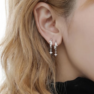 May include: Two silver-toned hoop earrings featuring a small, dangling star-shaped charm with a central clear stone. The hoops have a textured, irregular surface design and are worn in the earlobe, showcasing a metallic finish and delicate detail.