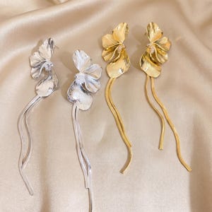 Puede incluir: Dos pares de pendientes largos colgantes, un par en plata y otro en oro. Cada pendiente presenta una flor texturizada de varios pétalos con dos cadenas finas y flexibles tipo serpiente que cuelgan del centro del diseño floral.