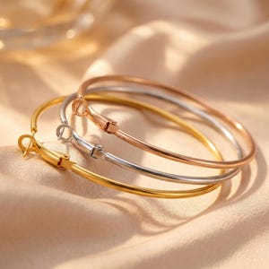 May include: Three hoop earrings in gold, silver, and rose gold. The earrings are displayed on a beige satin surface. Each earring has a simple, elegant design with a hinged closure.