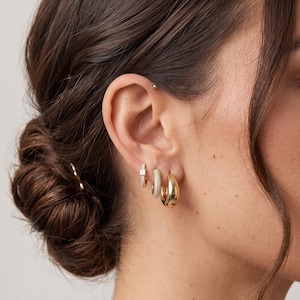 May include: Three gold-toned hoop earrings worn in a single earlobe. The smallest hoop features a single square-cut stone, the middle hoop is encrusted with small pavé stones, and the largest hoop is a smooth, polished, thick gold-toned band.
