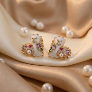 Pode incluir: Um par de brincos de botão dourados com um design em forma de coração. Cada brinco apresenta uma base de esmalte branco, adornada com pedras preciosas coloridas, incluindo roxo, vermelho e verde, e uma pérola. Os brincos estão sobre um fundo de cetim com pérolas espalhadas.
