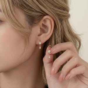 May include: A pair of rose gold hoop earrings featuring a small flower-shaped charm set with clear stones and a single dangling clear stone. The earrings are designed with a hinged closure mechanism, showcasing a delicate and refined metallic finish.
