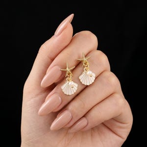 May include: Gold-toned earrings featuring a starfish and seashell design. The earrings have a white enamel seashell with a small pearl accent and a starfish charm. The earrings are dangling and have a post-back closure.